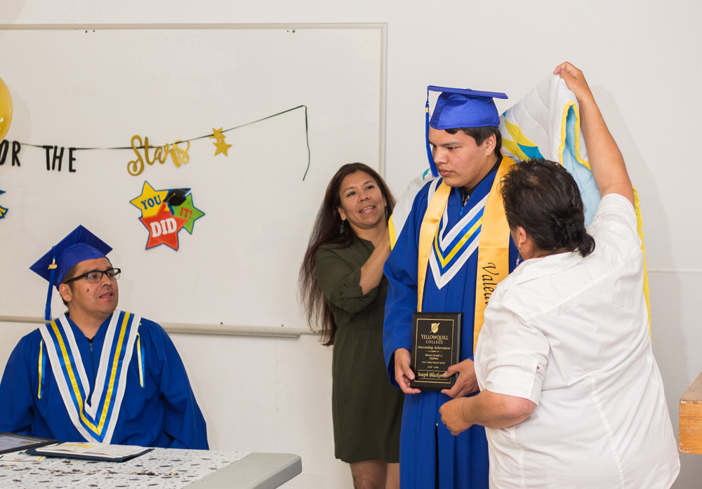 Sioux Valley Dakota Nation mature Grade 12 graduate Joseph Blacksmith is draped in a star blanket by Chief Jennifer Bone and student support worker Colleen Hall as fellow graduate Dustin Smoke watches at their graduation ceremony at the Wichozani Centre Friday. (Chelsea Kemp/The Brandon Sun)