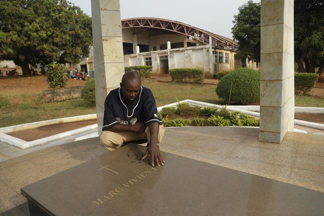 Assiga Pierre, nephew of the late Cameroonian soccer star Marc-Vivien Foé, touches the tombstone where he was buried near his abandoned soccer academy in Yaounde, Cameroon, Wednesday Feb. 2, 2022. The late Cameroon and Manchester City soccer star Marc-Vivien Foé had a dream to build a sports complex and school in his hometown of Yaounde. He never got to finish it after collapsing on a field while playing for his country in 2003 and dying of a heart condition at the age of 28. (AP Photo/Sunday Alamba)