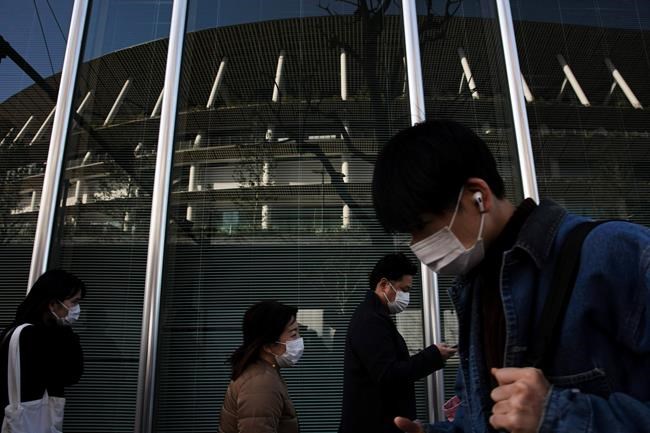People with masks walk past a building reflecting the New National Stadium, a venue for the opening and closing ceremonies at the Tokyo 2020 Olympics, Sunday, Feb. 23, 2020, in Tokyo. (AP Photo/Jae C. Hong)