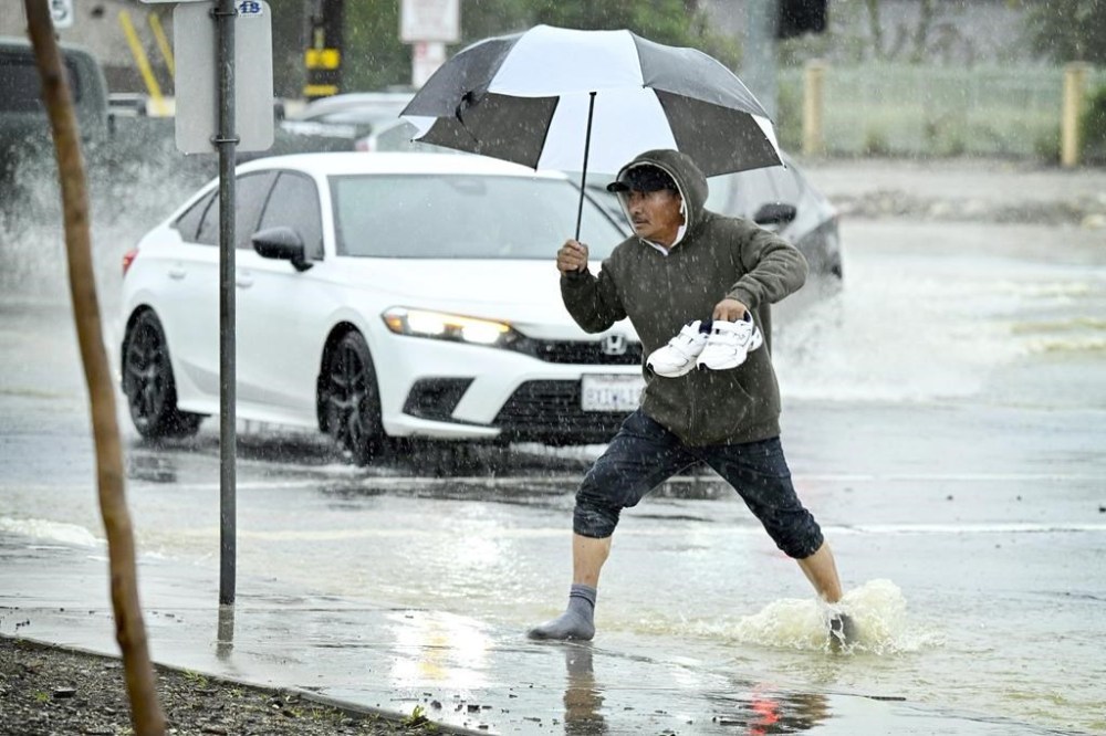 Braving the elements, Ismael Clazola carries his shoes as he crosses a flooded intersection at Foothill Boulevard and Cactus Avenue in Rialto, Calif., during a heavy rainstorm, Monday, Feb. 5, 2024. (Watchara Phomicinda/The Orange County Register via AP)