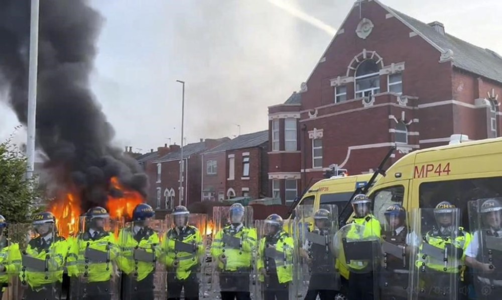 A police van, rear, burns as police line up after clashing with an unruly crowd, Tuesday, July 30, 2024, in Southport, northwest England, near where three girls were stabbed to death in a dance class the day before. The violence erupted shortly after a peaceful vigil was attended by hundreds in the center of Southport to mourn the 13 victims of the stabbings, including seven still in critical condition. (Pat Hurst/PA via AP)