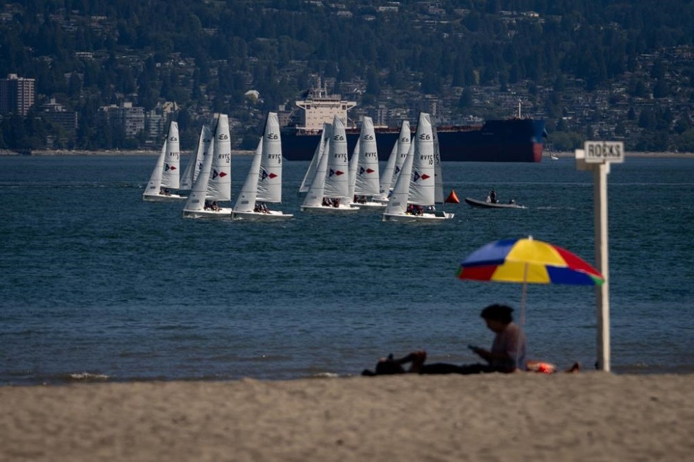 A person sits on the beach under a colourful umbrella as sail boats pass in Vancouver on July 7, 2024. THE CANADIAN PRESS/Ethan Cairns