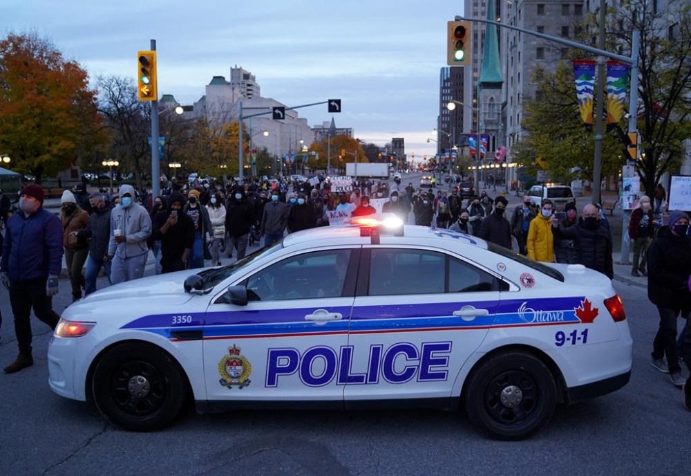 People pass a police officer in their car as they make their way along Elgin Street during a rally after an Ottawa police constable was found not guilty of manslaughter, aggravated assault and assault with a weapon in connection with the 2016 death of Abdirahman Abdi in Ottawa, on Oct. 20, 2020. THE CANADIAN PRESS/Justin Tang