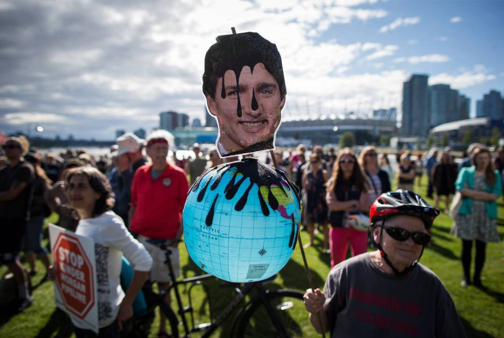 A protester holds a photo of Prime Minister Justin Trudeau and a representation of the globe covered in oil during a protest against the Kinder Morgan Trans Mountain Pipeline expansion in Vancouver, B.C., on May 29, 2018. (The Canadian Press files)