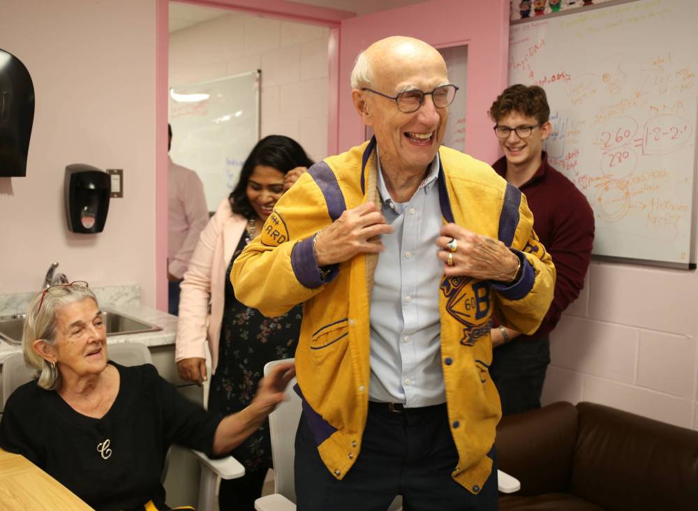 Gerald Butler smiles as he puts on the jacket he once wore as a student of Brandon College. Butler and his wife, Carmen (seated on the left), are longtime supporters of Brandon University and the Breast Cancer Cell and Molecular Research Laboratory. On Friday, a plaque in their honour was unveiled at the lab. (Michele McDougall/The Brandon Sun)