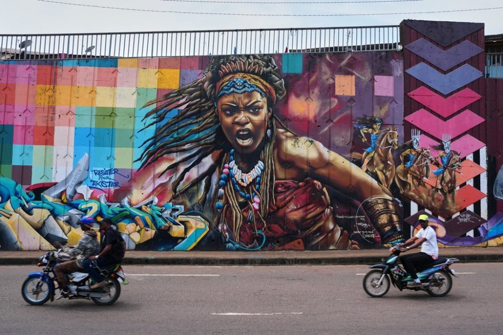 Motorcyclists ride past a mural depicting a powerful woman on a street wall in Conakry, Guinea, Sunday, Oct. 19, 2025. (AP Photo/Misper Apawu)