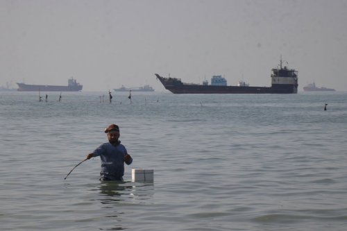 A man stands in the water, appearing to fish, as bulk carriers, cargo ships, and service vessels line the horizon in the Strait of Hormuz off Bandar Abbas, Iran, Monday, April 27, 2026.(Razieh Poudat/ISNA via AP)